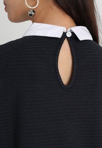 Svart texturerad tröja med en vit krage, med ett nyckelhålskärning bak, stängd med en knapp.