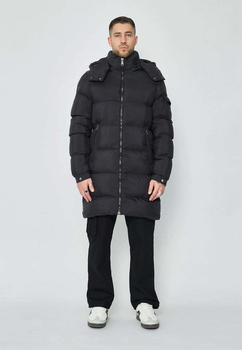 Manteau doudoune noir avec un col haut, fermeture éclair et poches latérales. Présente un design rembourré et couvre le milieu des cuisses. Porté avec un pantalon noir.