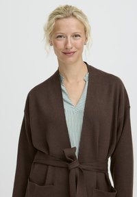 Donna bionda con occhi chiari, che indossa una camicia a righe blu chiaro sotto un cardigan marrone legato, sorridente su uno sfondo bianco neutro.
