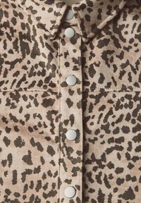 Chemise à imprimé léopard avec un tissu doux et texturé, fond tan avec des taches brunes foncées et des boutons-pression argentés sur le devant.