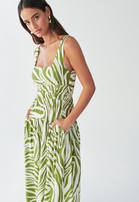 Robe maxi rayée verte et blanche avec un décolleté carré, une taille smockée et des poches latérales, fabriquée en tissu léger.