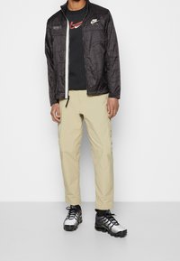 Nike mustän zip-up takki, millel on tekstureeritud muster, must t-särk punase logoga, beežid püksid ja hõbedased spordijalatsid mustade aktsentidega.