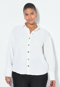 Una mujer con el pelo corto sonríe, vestida con una camisa blanca abotonada, con botones plateados, y pantalones negros contra un fondo blanco.