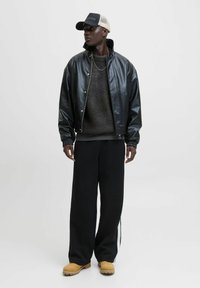 Mand iført sort læderjakke, mørk sweater, brede sorte bukser med hvid stripe, brune støvler og en sort og beige baseballcap, der kigger til siden.