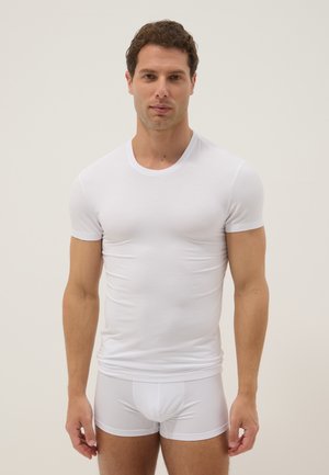 Hombre con cabello corto y castaño, vistiendo una camiseta blanca ajustada de manga corta y calzoncillos bóxer blancos, de pie frente a un fondo liso.