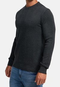Ciemnoszary sweter wykonany na drutach z wzorem jodełki, długimi rękawami i okrągłym dekoltem, wykończony prążkowanymi ściągaczami na mankietach i dole. W połączeniu z niebieskimi dżinsami.