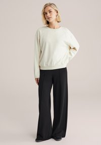 Hanorac crem deschis cu croială lejeră, manșete și tiv cu rebră. Asortat cu pantaloni negri cu picioare largi și încălțăminte închisă la culoare. Fundal neutru.