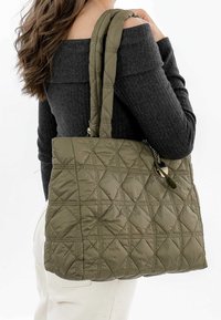 Sac fourre-tout matelassé vert olive avec des anses en tissu doubles et des accents en métal argenté. Présente un motif en losanges et une surface douce et texturée.