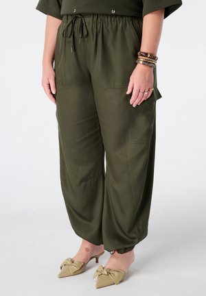 Persoon draagt olijfgroene wijde broek met koord in de taille, beige mules met hak en strikjes, en meerdere armbanden aan de rechterpols.