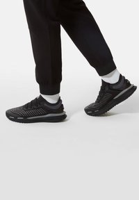 Svarta sportskor med en texturerad ovandel, prydda med ljusgrå detaljer och slitstarka sula, bärs tillsammans med svarta joggers och vita strumpor.