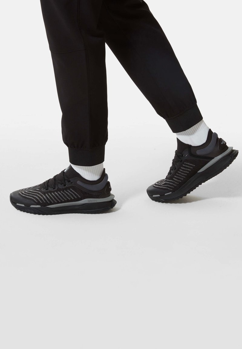 Svarta sportskor med en texturerad ovandel, prydda med ljusgrå detaljer och slitstarka sula, bärs tillsammans med svarta joggers och vita strumpor.