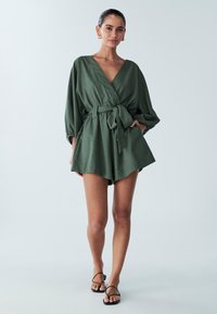 Green linen wrap romper with a v-neck, cinched waist, and wide sleeves. Features a tie belt and relaxed shorts. Black strappy sandals.