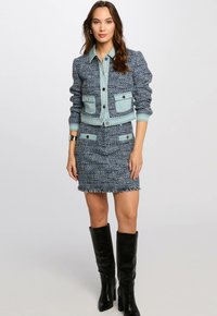 Blauwe tweedjas met lichtblauwe denimaccenten en knopen, gecombineerd met een strakke blauwe tweed minirok met franjerand. Zwarte kniehoge laarzen.