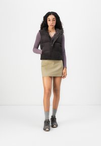 Woman standing wearing a black puffer vest, purple long-sleeve top, beige skirt, gray socks, and dark hiking shoes against a white background.