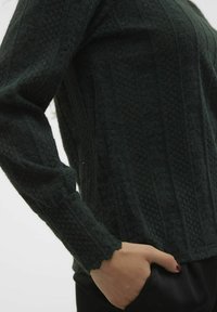 Mörkgrön stickad tröja med texturerade mönster, långa ärmar, vågiga manschetter och en avslappnad passform med en hand i fickan.