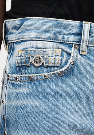 Džinsai su sidabriniais kaiščiais ir mažyte priekinės kišenės detalė, kurioje ant šviesiai mėlynos medžiagos yra išraiškinga sidabrinė Medūzos galvos emblema.