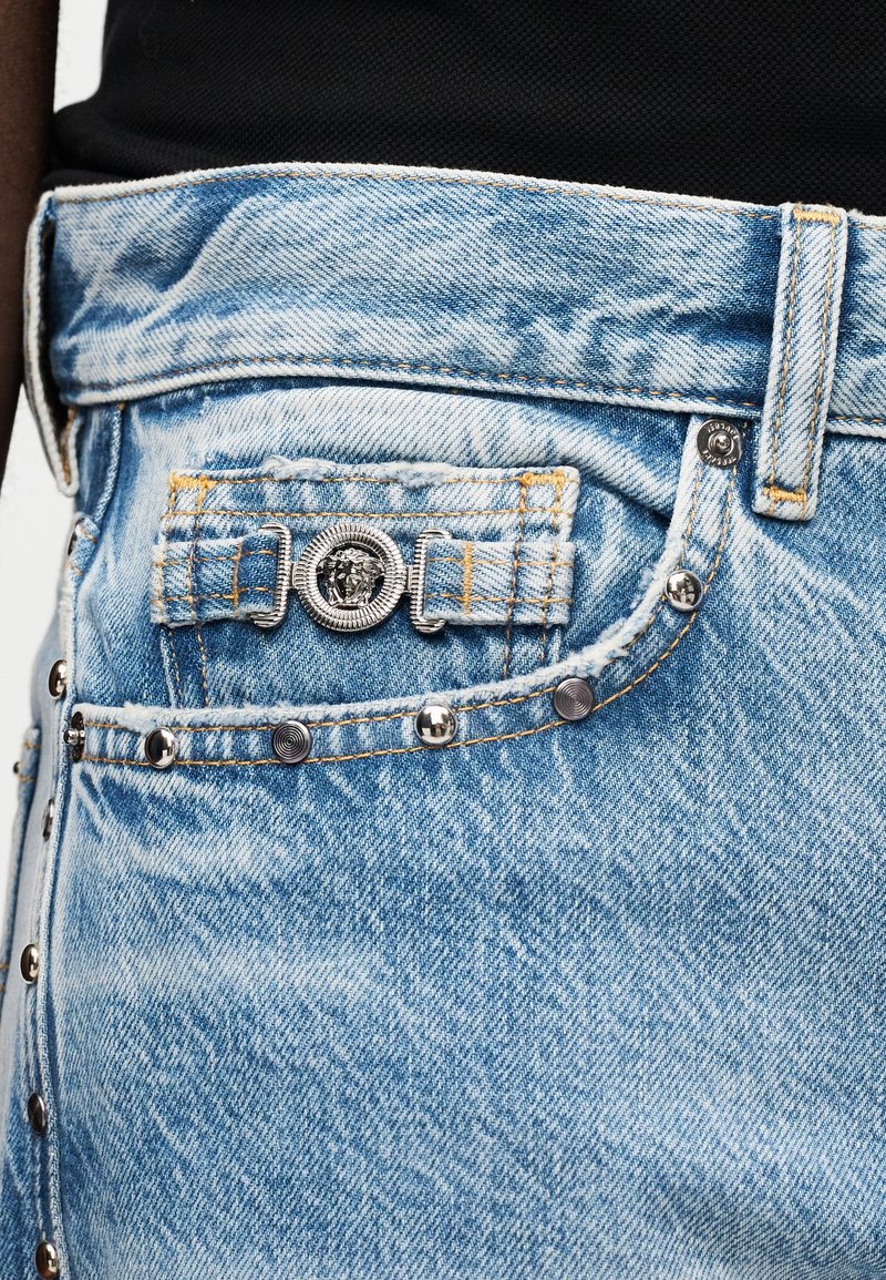 Jean en denim avec des clous en argent et une petite poche avant ornée d'un emblème détaillé de tête de Méduse en argent sur un tissu bleu clair.