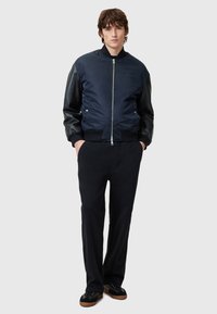 Navy bomber jacket with a smooth fabric body, black leather sleeves, ribbed cuffs, and a zipped front, paired with dark trousers and shoes.