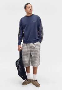 Tmavomodrá mikina s kontrastnými bočnými pruhmi, sivé kárované šortky, hnedé topánky a čierna taška na rameno s bielym logom.