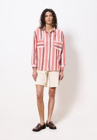 Rot-weiß gestreiftes Langarmhemd mit zwei Brusttaschen, kombiniert mit cremefarbenen Shorts und braunen Mokassins, vor einem weißen Hintergrund stehend.