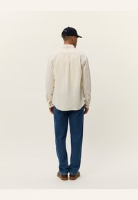 Light-colored long-sleeve shirt with button cuffs, textured fabric, and a back pleat. Paired with blue denim jeans and a dark cap.