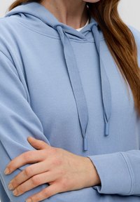 Sudadera azul clara hecha de tela suave, con capucha ajustable, puños acanalados y una textura lisa. En primer plano se ve el brazo cruzado.