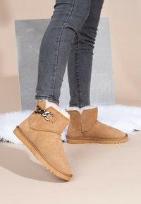 Botines de ante en tono beige con forro de piel sintética y detalle de cadena en el costado, con punta redonda y suela de goma marrón.