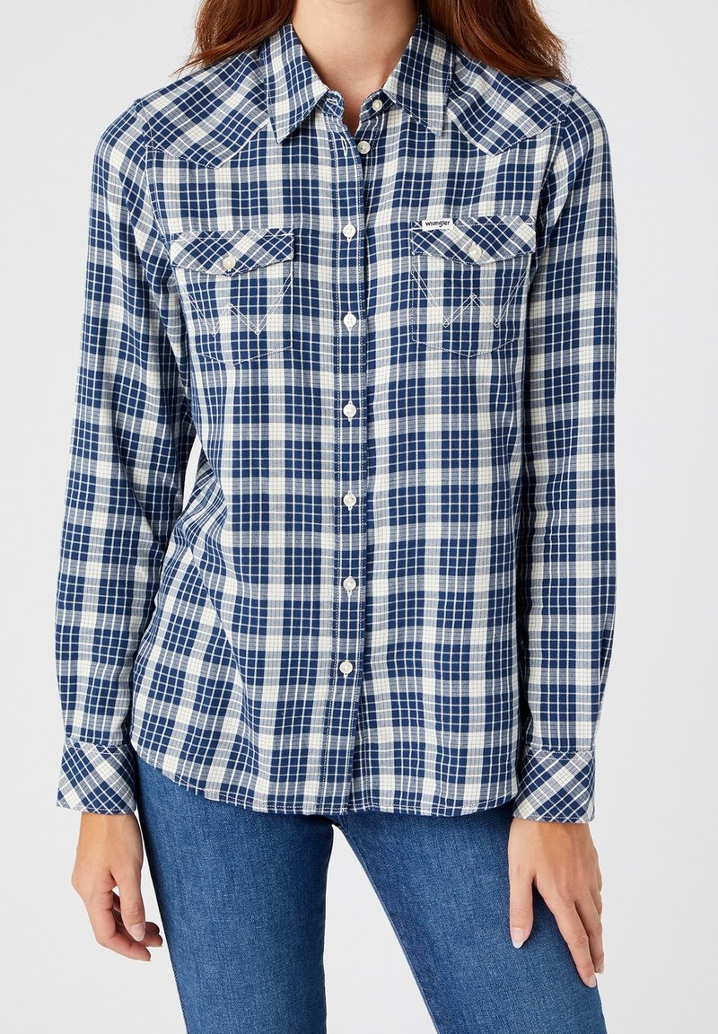 Femme portant une chemise à carreaux bleue et blanche avec des poches poitrine à boutons, associée à un jean bleu, debout devant un fond uni.