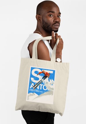 Uomo con barba e orologio tiene una borsa tote beige con l'immagine di uno sciatore con giacca rossa su una pista innevata con cielo azzurro sullo sfondo.