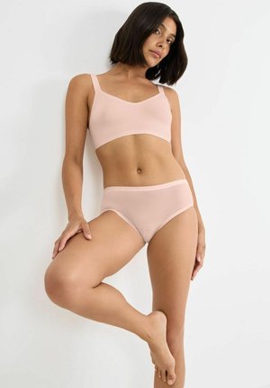 Woman with short dark hair posing barefoot against white wall, wearing light pink ribbed bralette and matching high-waist underwear.