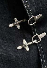 Δύο ασημένια κουμπώματα με άγκιστρα και μπαρ προσαρτημένα σε σκούρο μπλε ντένιμ ύφασμα με ορατή λευκή ραφή.