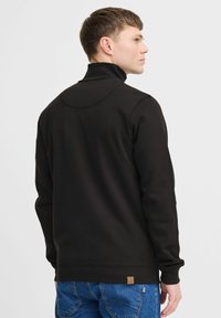 Black sweatshirt with high collar, soft texture, ribbed cuffs, and hem. Features a small brand label on the lower left side.