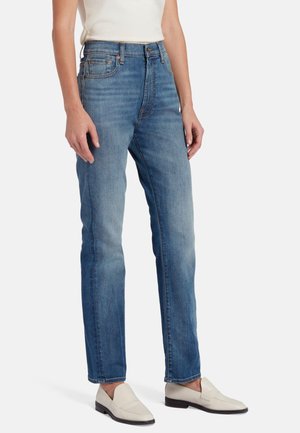 Jean taille haute bleu clair avec une coupe droite, comportant cinq poches et une couture visible. Associé à des mocassins blancs.