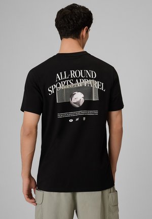 Mężczyzna w czarnej koszulce z grafiką bramki piłkarskiej i białym napisem "All-Round Sports Apparel" na plecach, do tego jasne szorty.