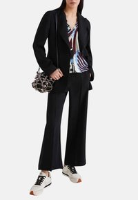 Giacca nera su misura sopra una blusa a fantasia, abbinata a pantaloni a gamba larga e sneakers bianche. Presenta una borsa di perline.
