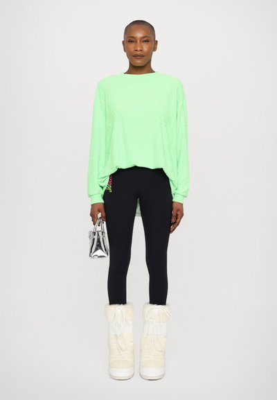 Felpa a maniche lunghe verde neon con logo in rilievo, leggings neri, stivali bianchi foderati in pelliccia e una piccola borsa a tracolla argentata.