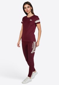 Burgunderfarbener Sportanzug mit einem taillierten T-Shirt mit cremfarbenen Streifen und einem Löwenlogo, kombiniert mit Leggings, die den Markennamen in Weiß anzeigen.