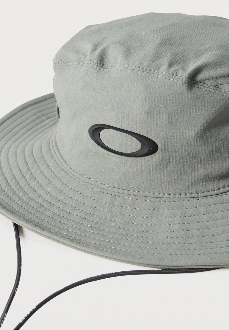 Chapeau bob gris clair avec un large bord, une lanière de menton réglable et un logo ovale noir au centre avant.