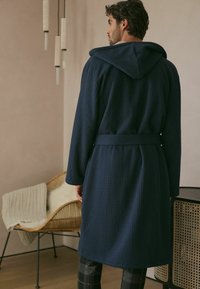 Tumedat sinine vahvline tekstuuriga hommikumantel kapuutsiga, vöökohaga ja pikkade varrukatega, nähtud seljaga minimalistlikus sisevõinangus.