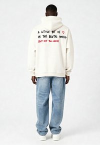 Krems krūšturis ar kapuci, uz muguras ir melns un sarkans teksts. Apvienots ar gaiši zilo platā kāju džinsu un baltām sporta kurpēm.