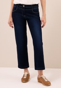 Persona vistiendo jeans rectos de color azul oscuro y zapatillas marrones con cordones blancos, de pie frente a un fondo beige liso.