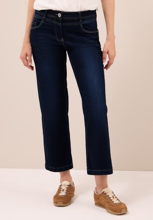 Persoon in donkerblauwe rechte jeans en bruine sneakers met witte veters, staande tegen een effen beige achtergrond.