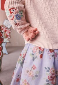 Child wearing a pink knitted sweater with embroidered flowers on the sleeve and a light purple floral skirt.