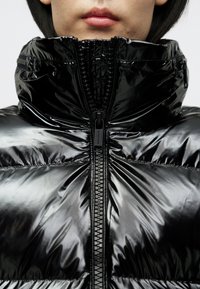 Κοντινό πλάνο ενός ατόμου που φοράει μια γυαλιστερή μαύρη μπουφάν με ψηλό γιακά κλεισμένο ως τον πηγούνι.