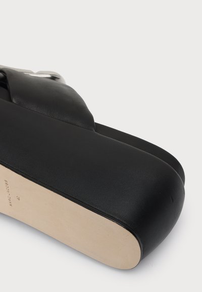 Gros plan sur une sandale plateforme noire avec une épaisse semelle et une bride en cuir croisée, marque Marc Jacobs visible sur la semelle.