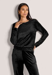 Vrouw met lang donker haar, gekleed in gouden oorbellen en een zwart fluwelen top met luipaardprint met lange mouwen, en bijpassende broek, poserend tegen een lichte muur.