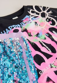Schwarzes T-Shirt mit farbenfrohem grafischem Text und Blumenmuster, geschichtet mit einem lila Stoff, der mit blauen Pailletten verziert ist.