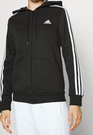Persoon draagt een zwarte Adidas zip-up hoodie met witte 3-strepen details op de mouwen en een wit Adidas-logo op de borst.