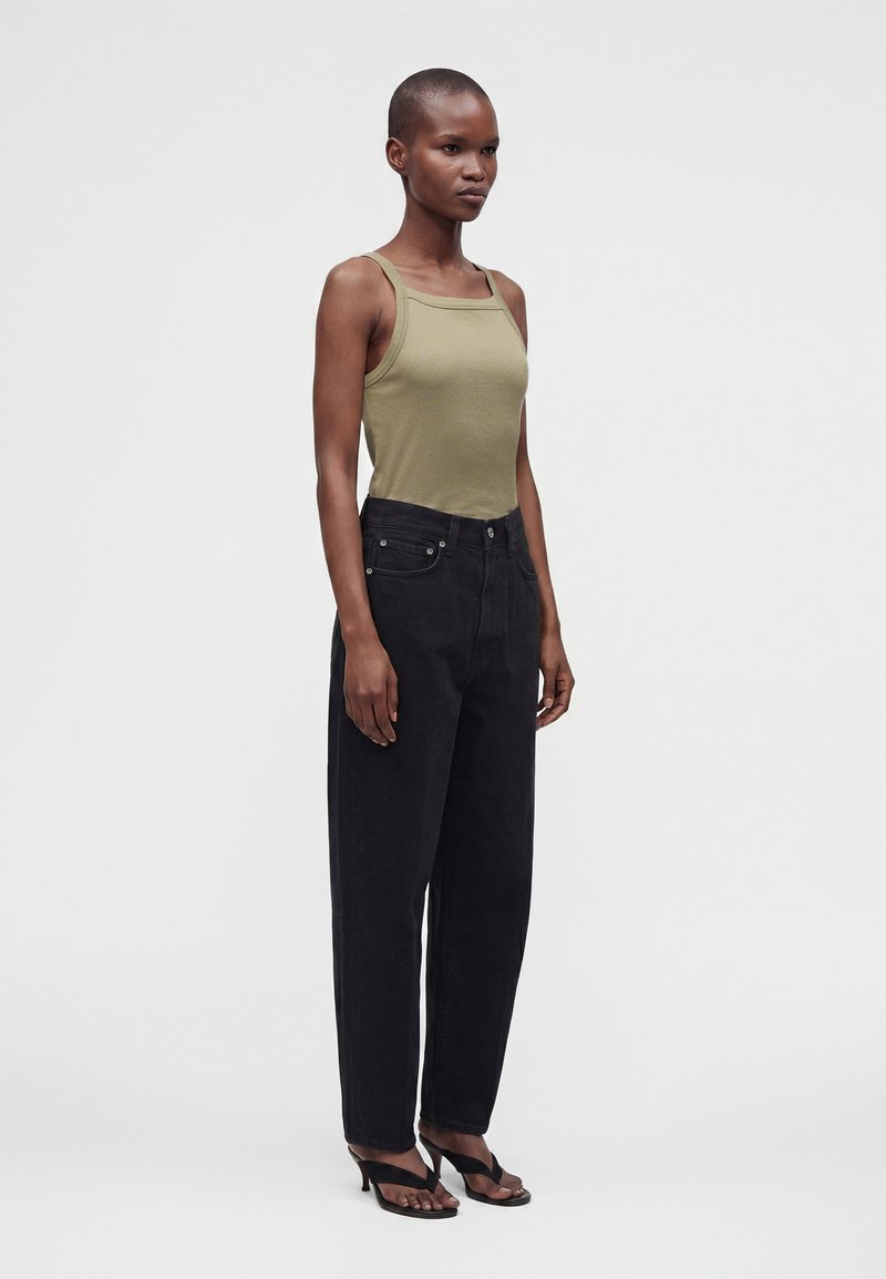 Femme debout portant un débardeur vert olive, un jean noir taille haute et des sandales à talons noires sur un fond gris uni.