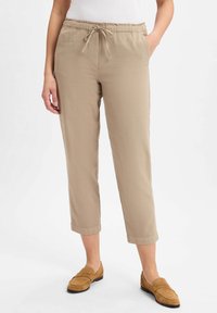 Femme portant un pantalon beige court à cordon de serrage et des mocassins en daim beige, se tenant avec une main dans la poche contre un fond blanc.